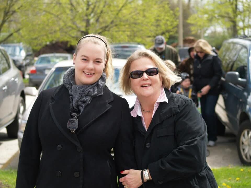 Two women in black coats smiling together outdoors, with cars and trees in the background.