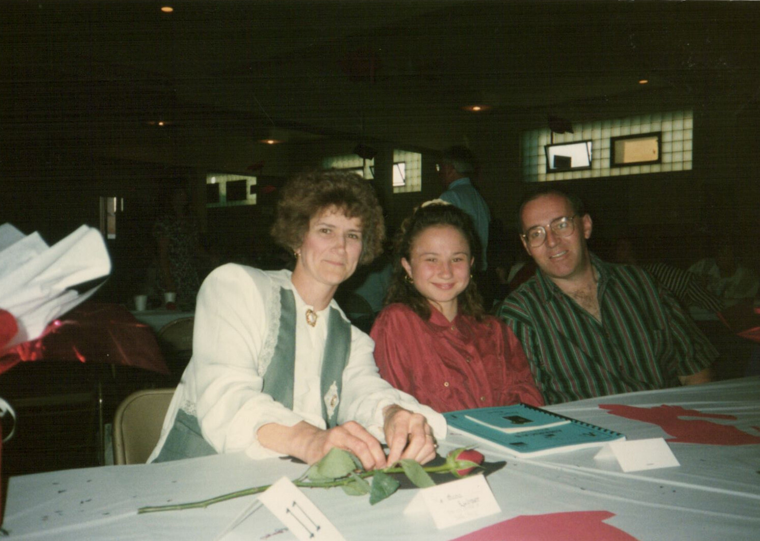 Three people pose for a photo at a table during an event; a woman with curly hair on the left, a smiling girl in a red blouse in the middle, and a man in a striped shirt on the right sit together inside a hall with name cards and a rose on the table.