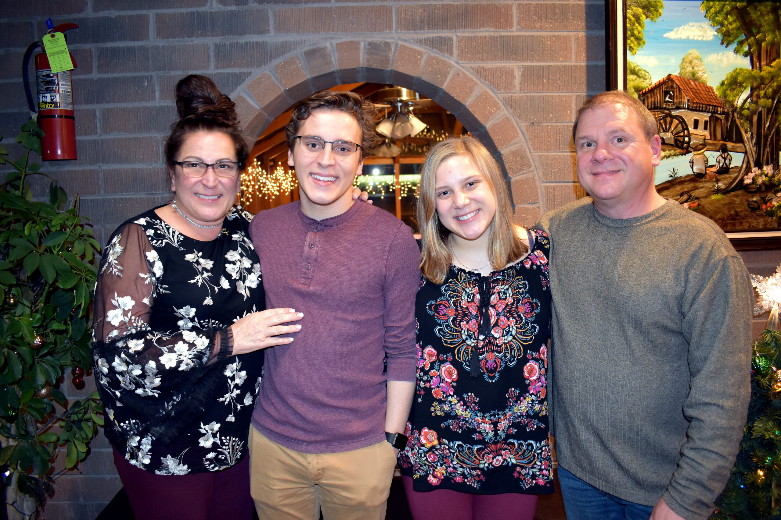 Four adults posing together indoors in front of a brick arch, smiling at the camera. Festive lights glow in the background.