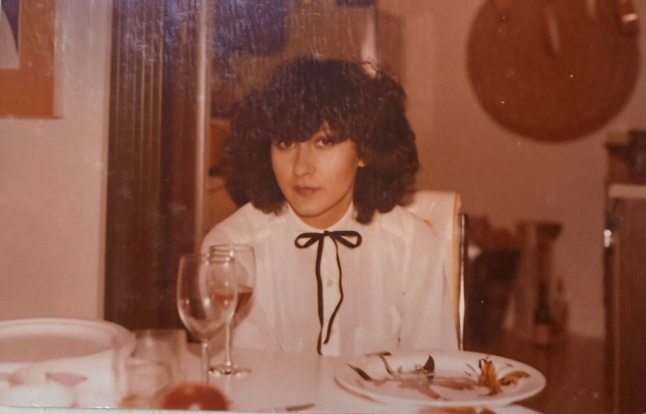 Woman with dark curly hair in a white blouse with a black ribbon sits at a dining table with a wine glass and plate of food; warm background with a guitar on the wall.
