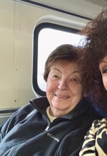 Smiling elderly woman seated by a window inside a vehicle, with another person partially visible beside her.