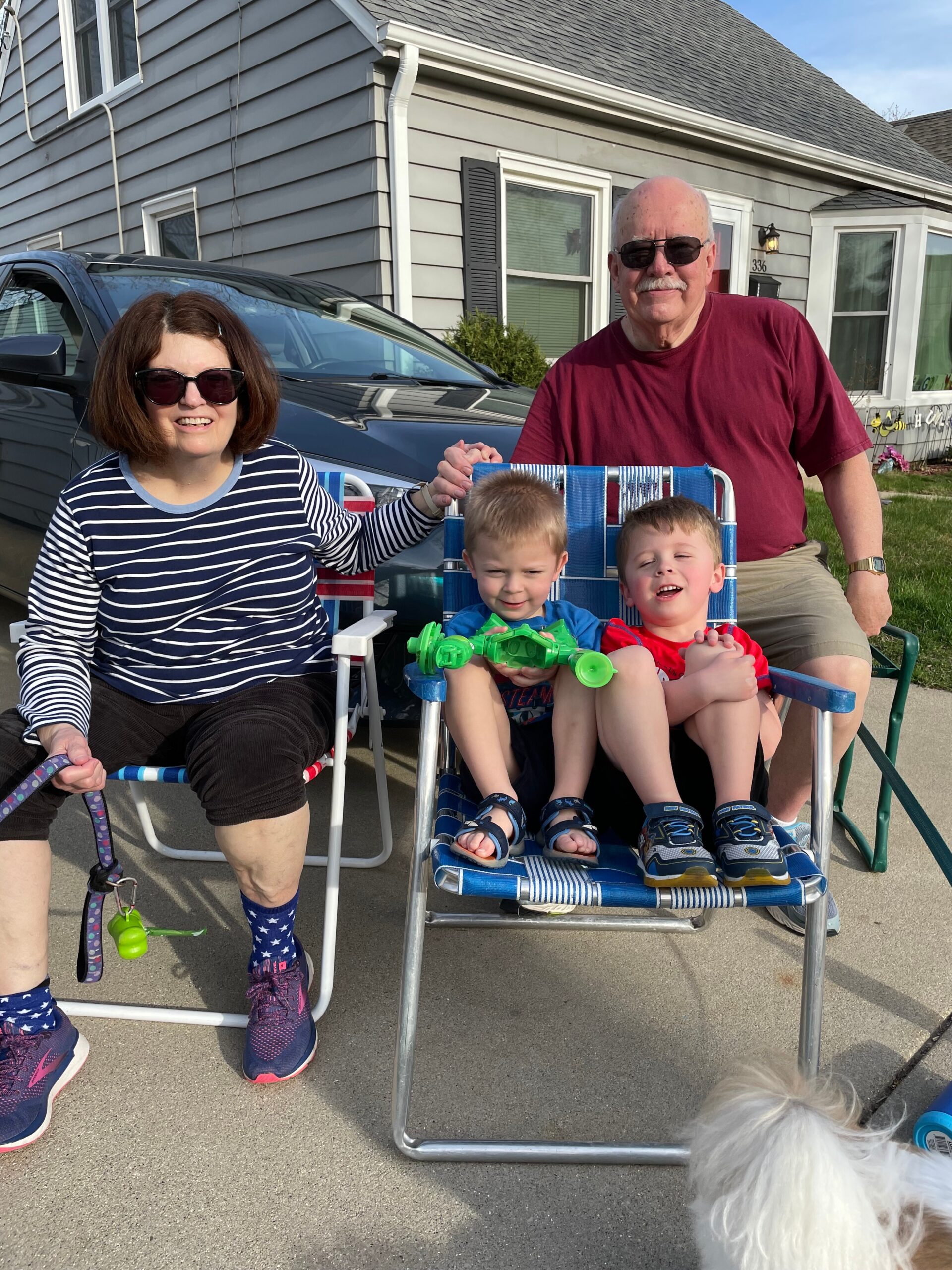A woman and an older man posing with two young boys sitting in a blue wagon outside a suburban house and car in the background.