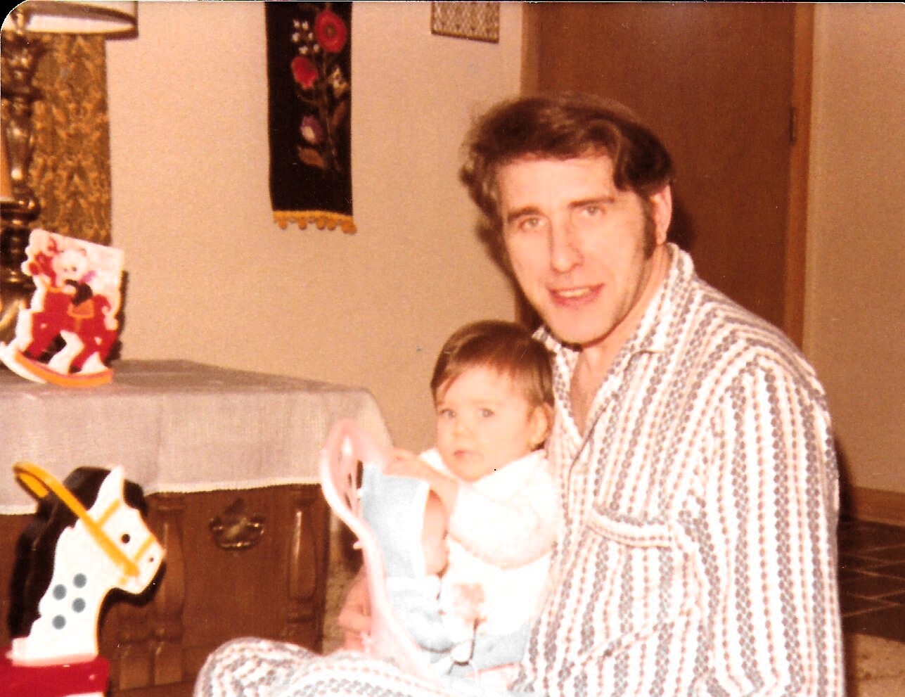 Man in striped pajama top with a baby on his lap, sitting in a living room with a rocking horse toy nearby and holiday decorations on a table behind them.