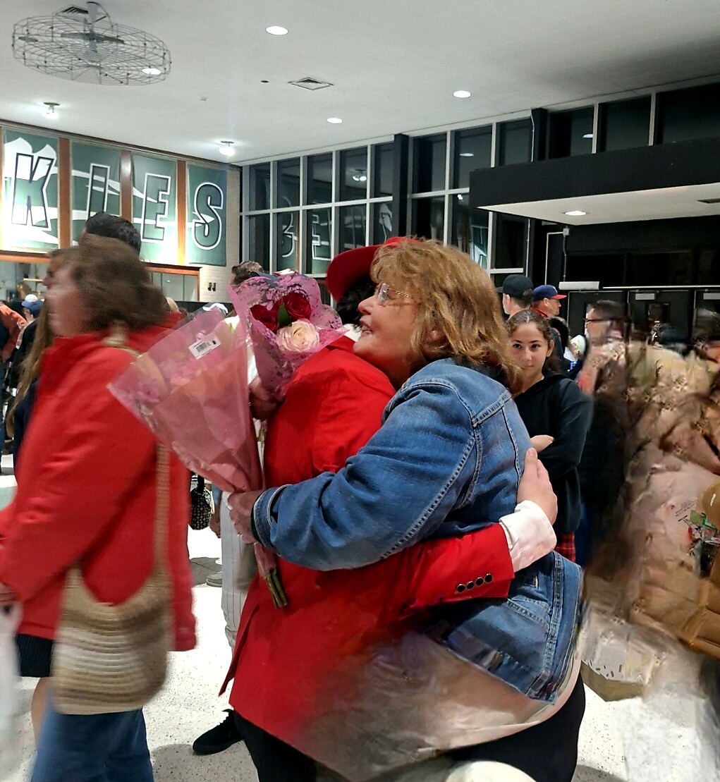 Two people share a hug in a busy school hallway, one holding a bouquet of pink flowers.