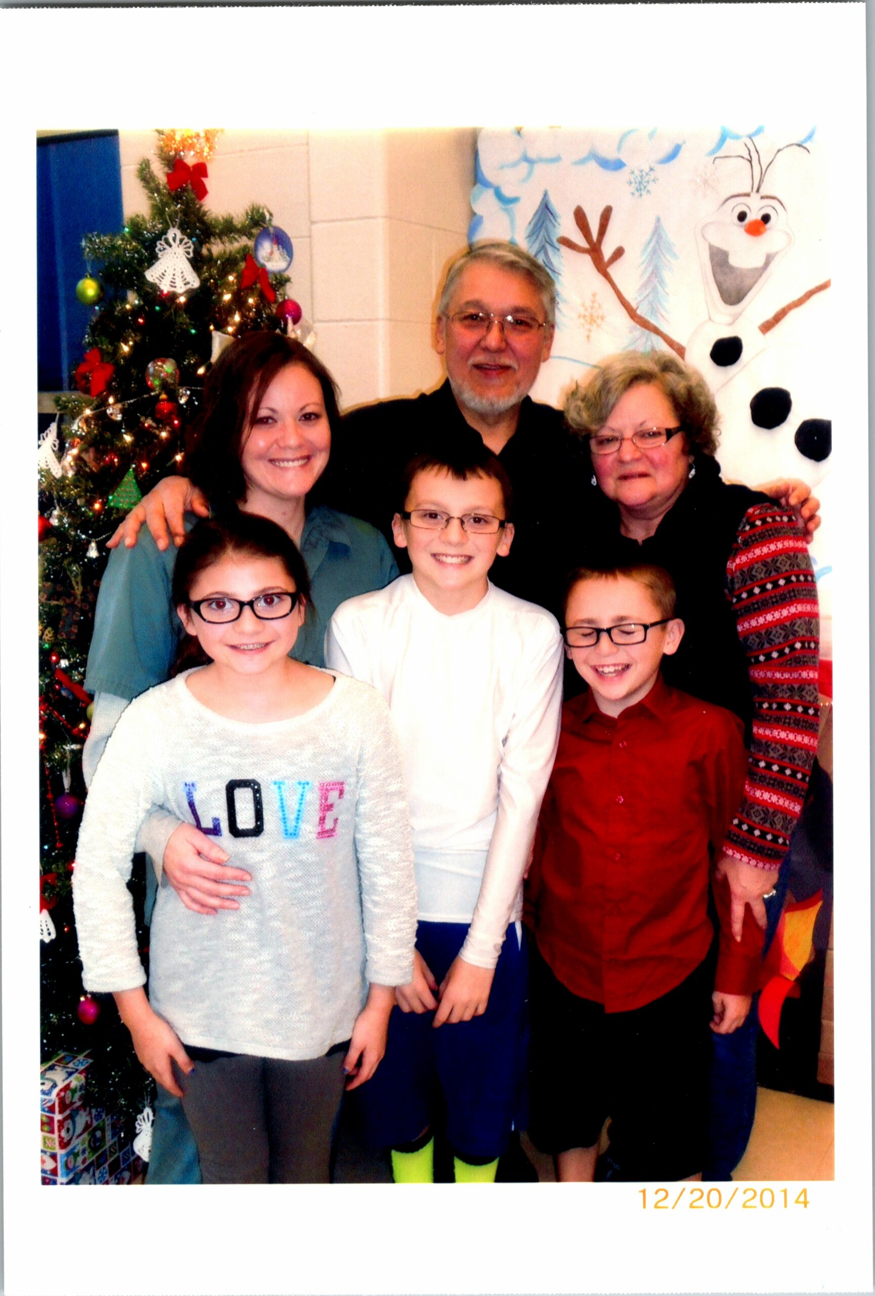 A family group photo indoors beside a decorated Christmas tree and a winter Olaf mural, with six smiling children and adults posing together.