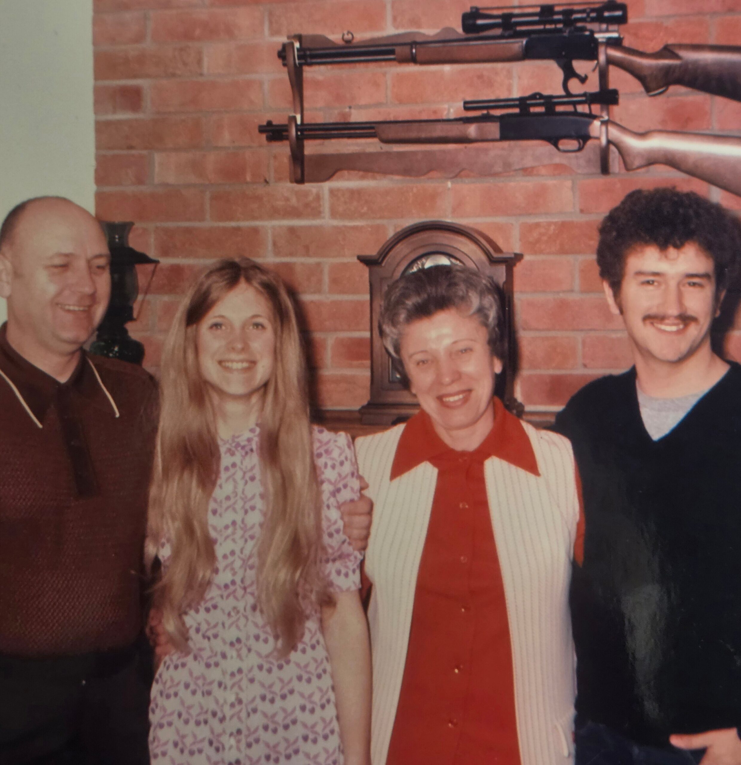 Group of smiling adults posing for a photo in front of a brick wall with two mounted rifles visible above them in the background.
