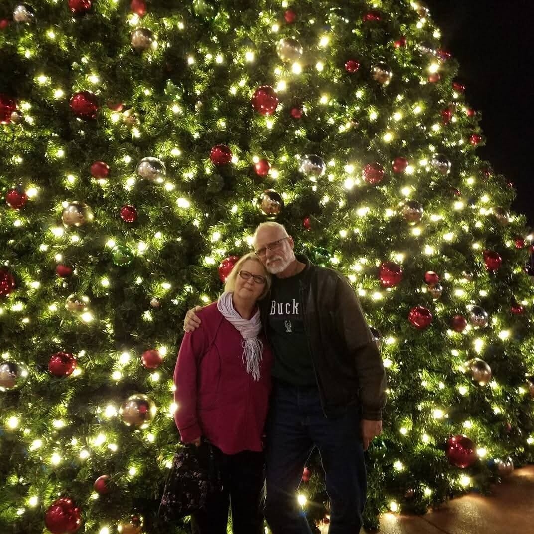 Older couple posing together in front of a large, illuminated Christmas tree with red and silver ornaments.