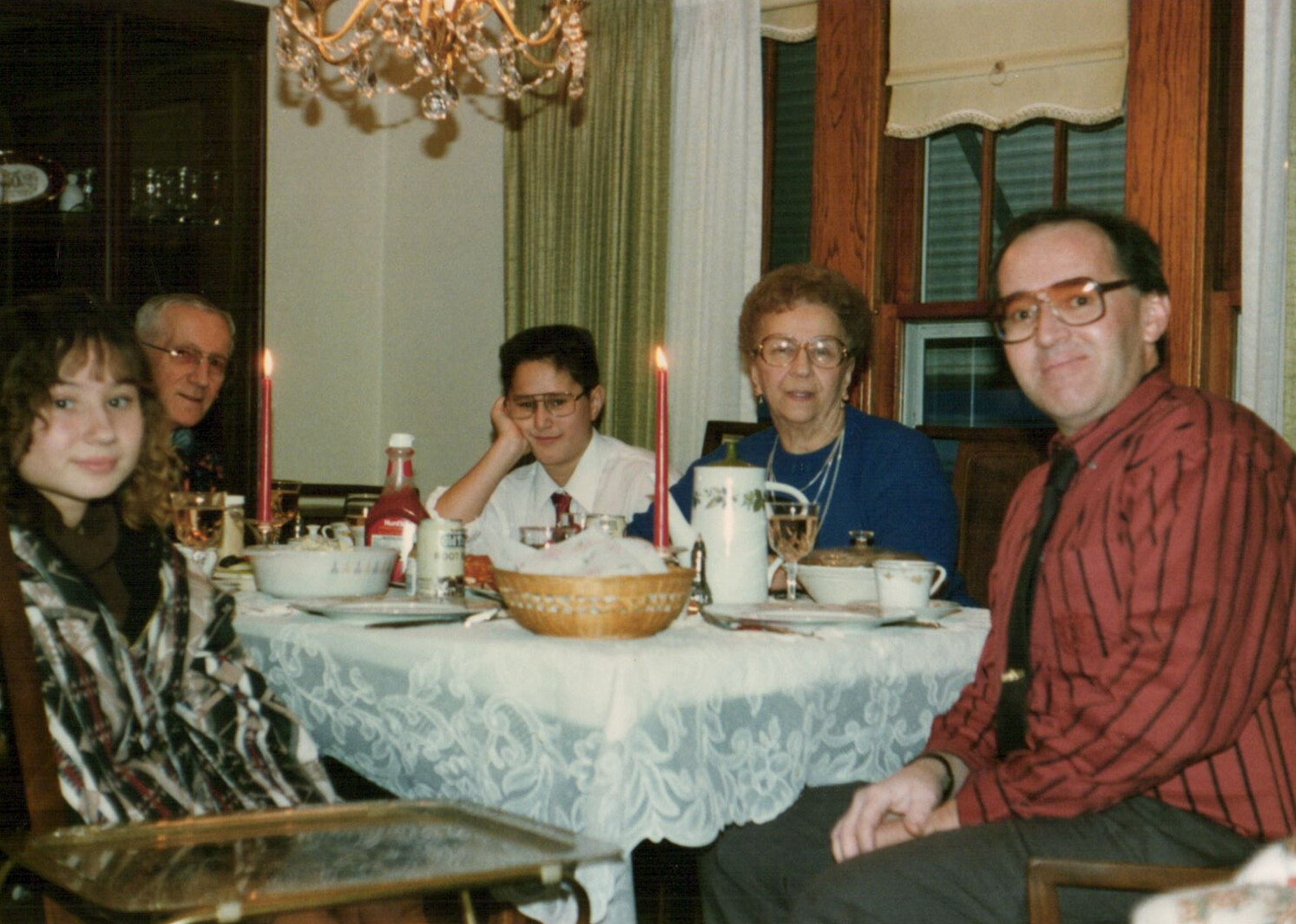 Family members sit around a lace-covered dining table with candles and food, posing for a photo at a meal.