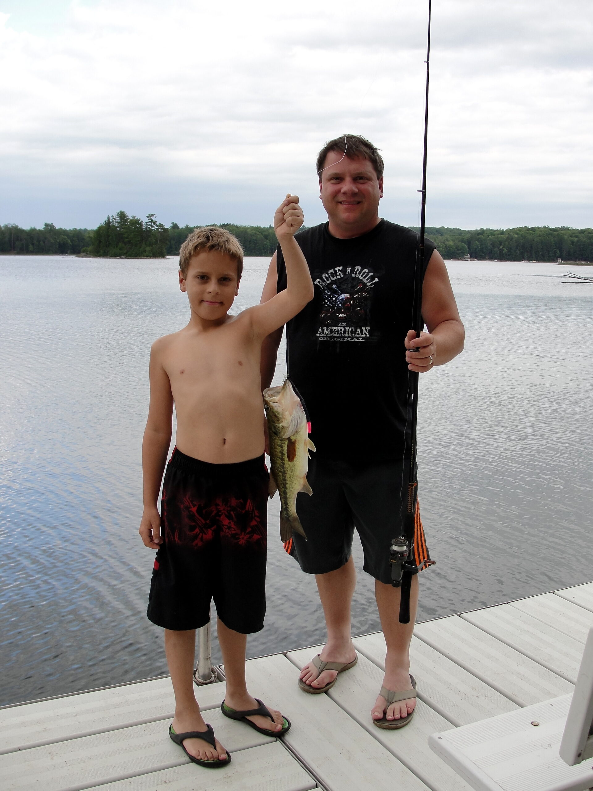 Two people stand on a wooden dock by a lake; the adult holds a fishing rod and a large fish while the boy stands beside him.