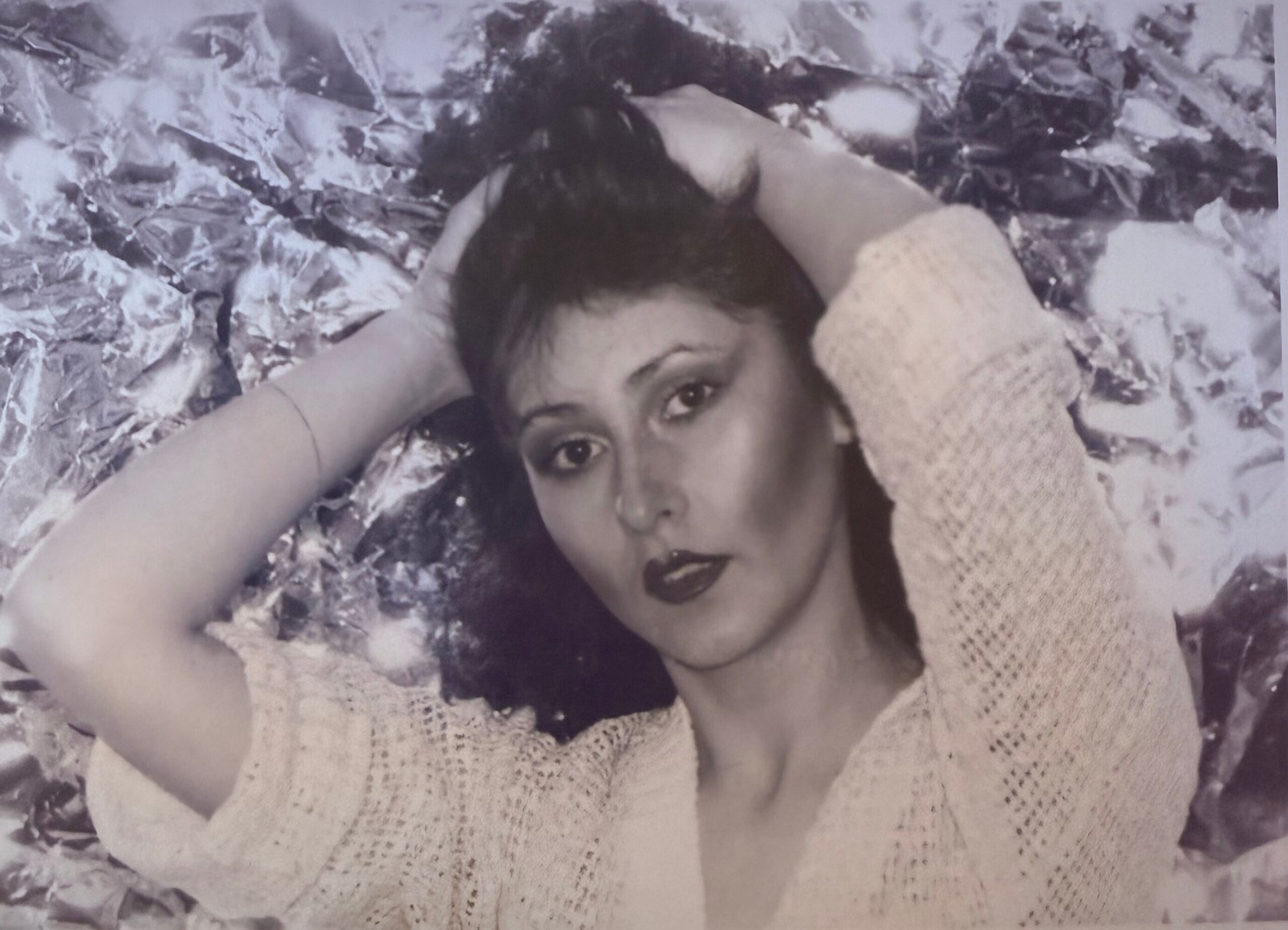 Young woman lying on a crumpled metallic foil background, head resting on arms, wearing a light knit sweater.