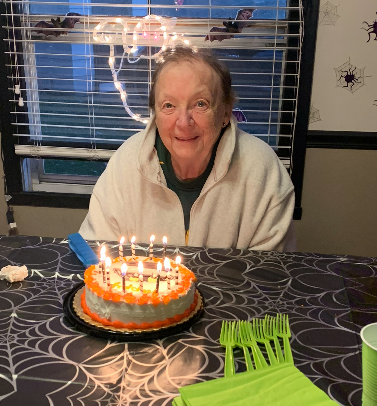 Smiling elderly woman in a light jacket sits at a table with a two-layer birthday cake and lit candles in front of her, Halloween decor in the background.