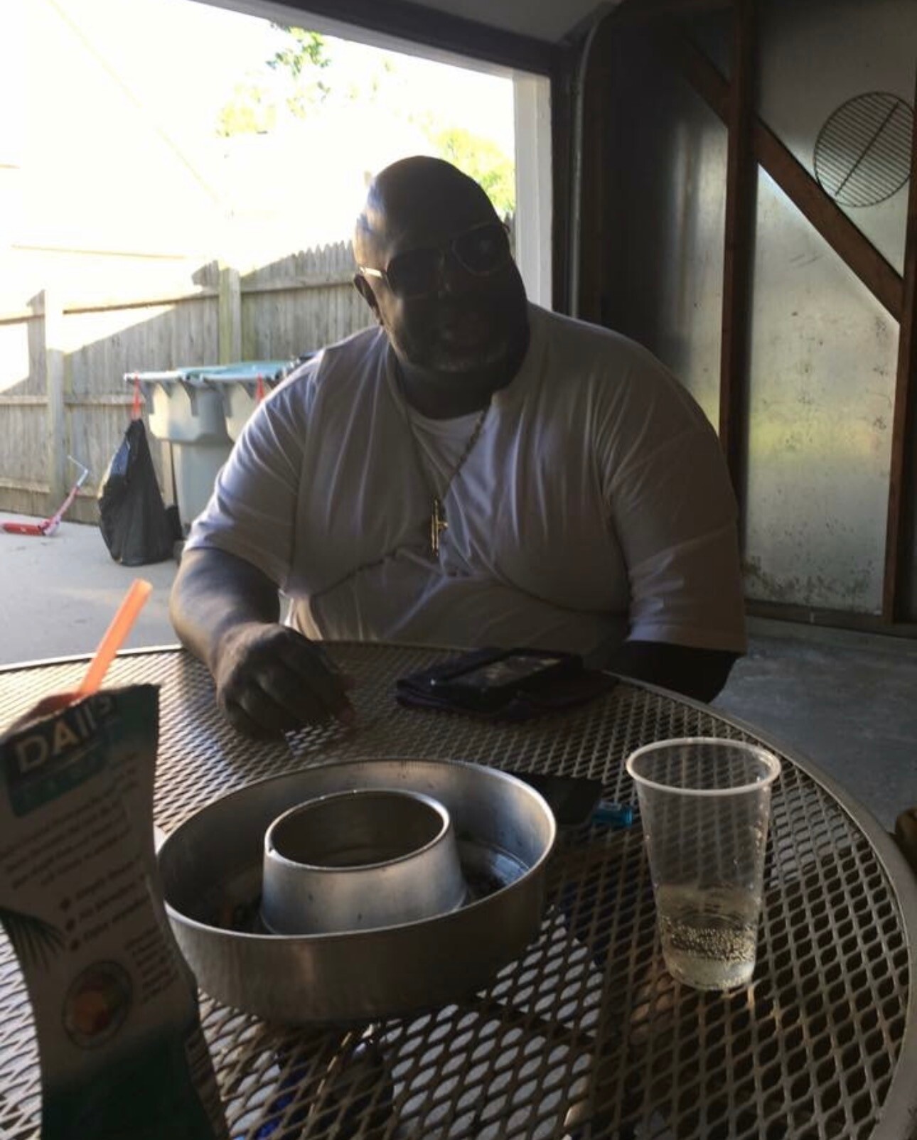 Man in a white T-shirt and sunglasses sits at a round metal patio table, with a Bundt pan and baking gear in front, outdoors in a backyard.