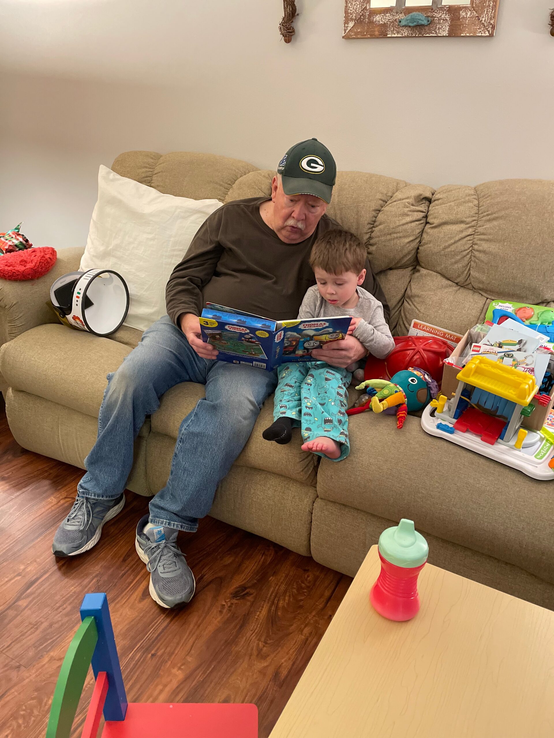An elderly man wearing a green Packers cap sits on a beige sofa, reading a colorful children’s book with a young boy beside him.