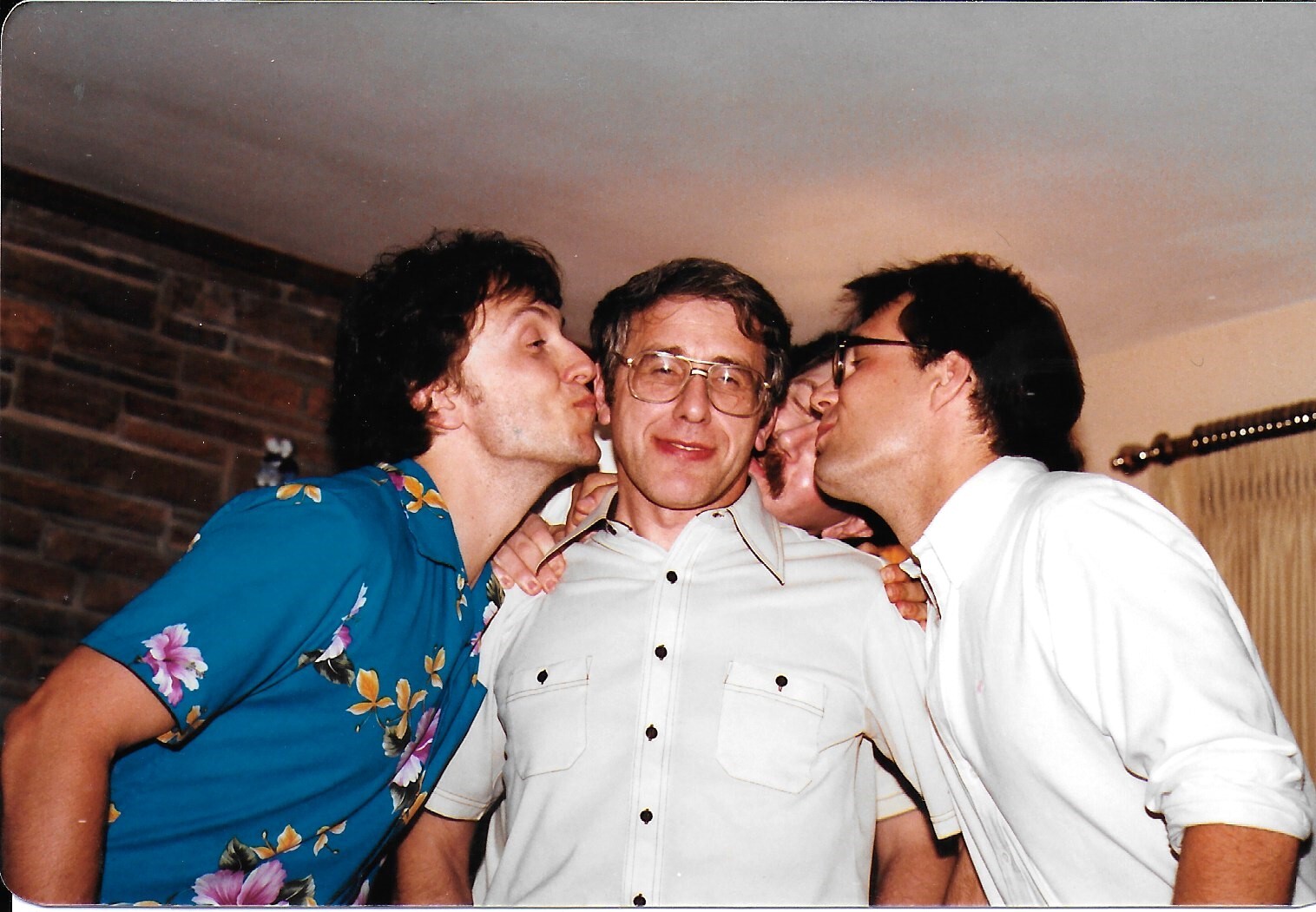 Three men at a party; two friends kiss the man in the middle on the cheeks while he smiles.