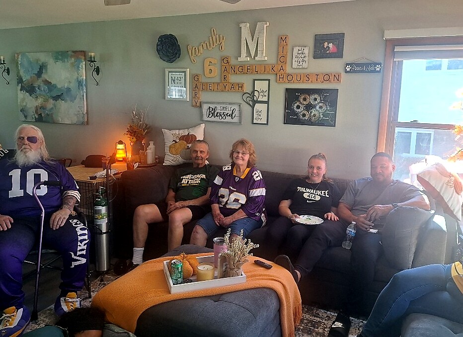 Six adults sit on a living room sofa wearing Minnesota Vikings jerseys; a coffee table with drinks and snacks sits in front, wall art behind them.