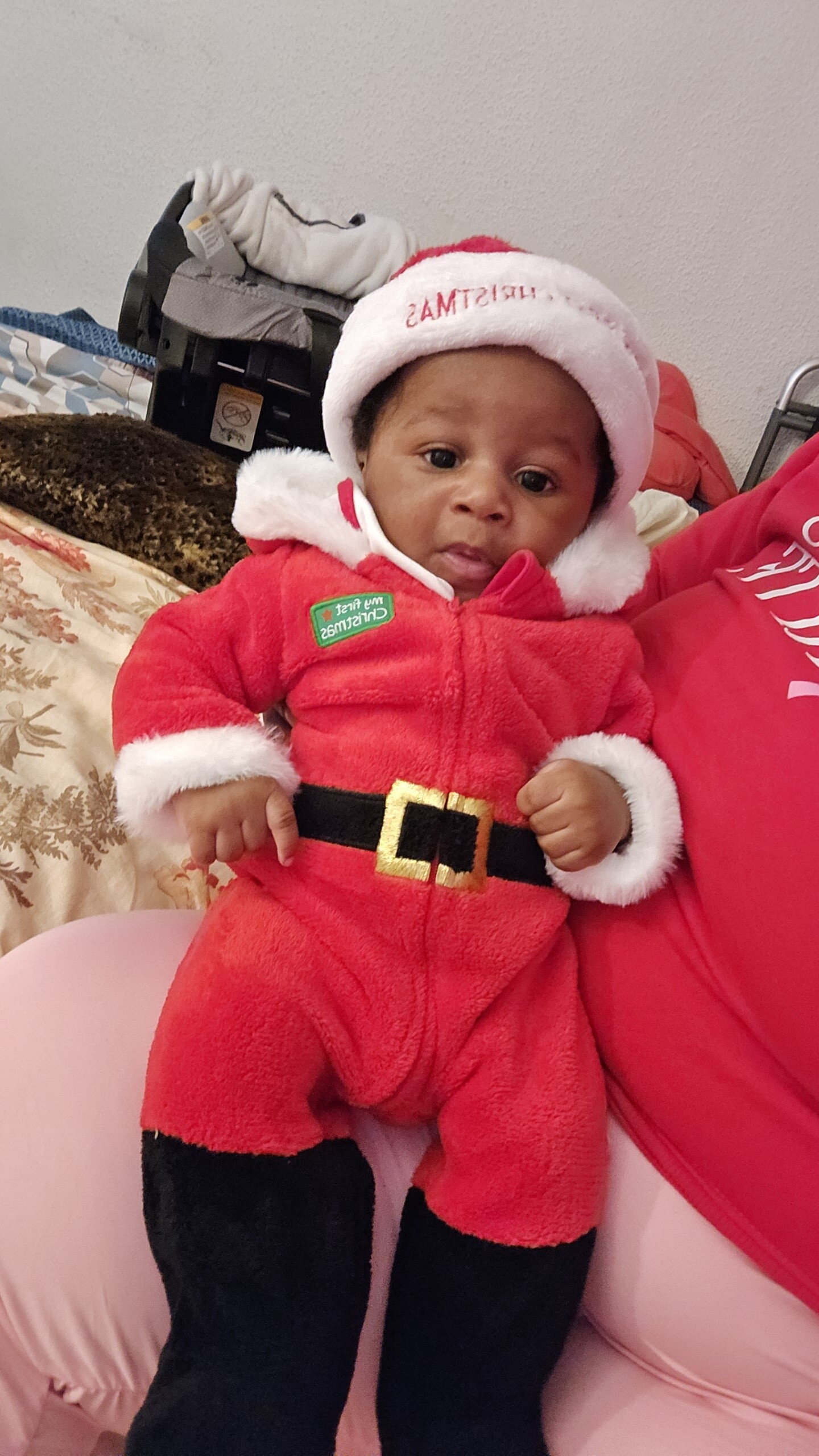 Baby wearing a fluffy red Santa suit and hat, sitting on a pink chair and looking at the camera.