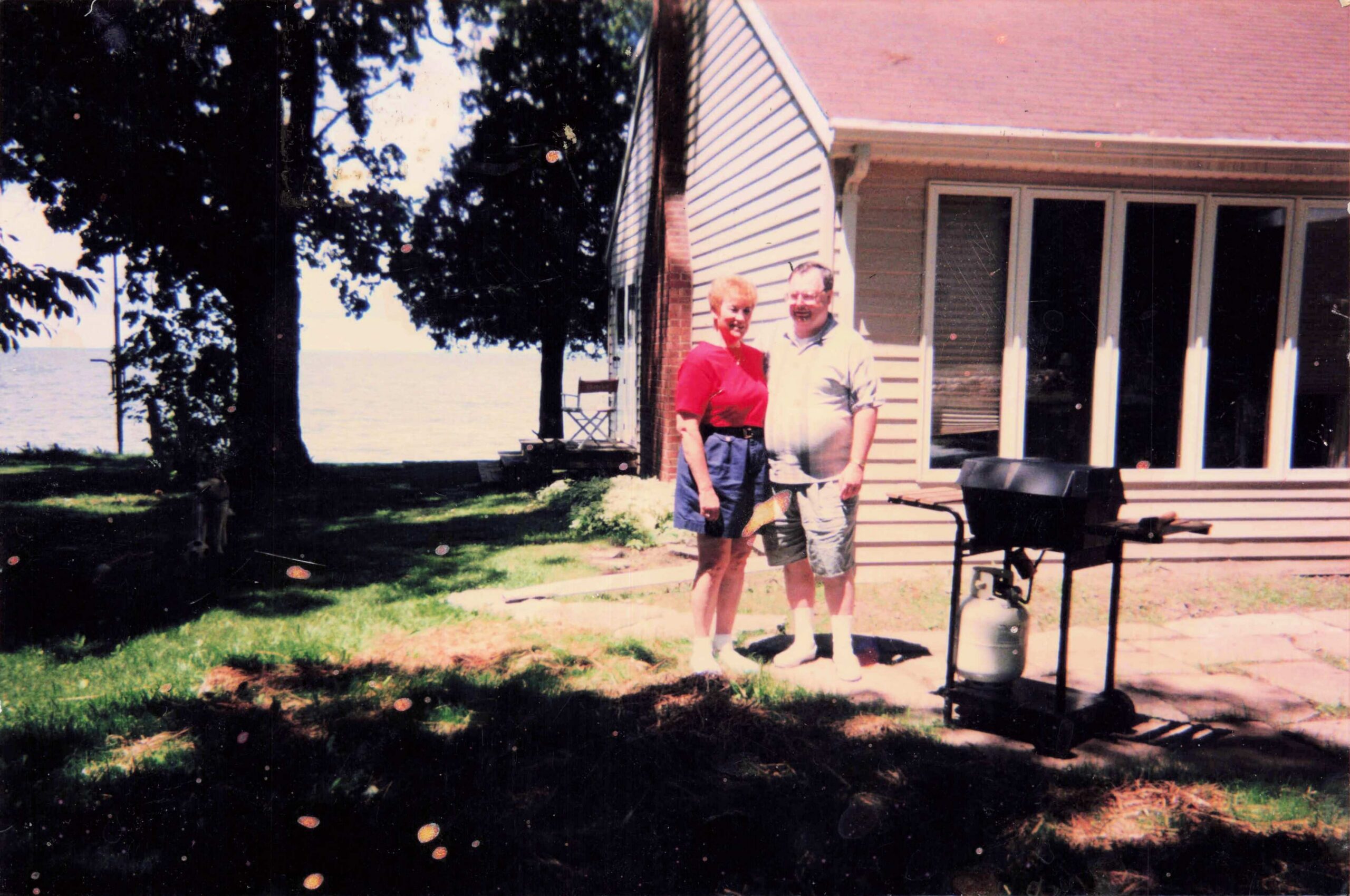 Couple posing on a lawn by a lakeside house, with trees and a grill in the foreground.