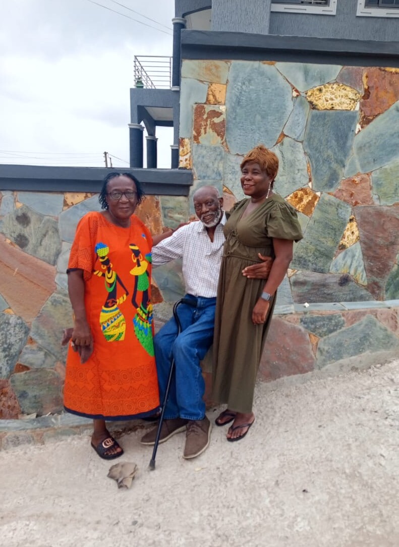 Three adults pose together outdoors beside a stone wall; an elderly man with a cane stands between two women, one in an orange dress and the other in a green dress.
