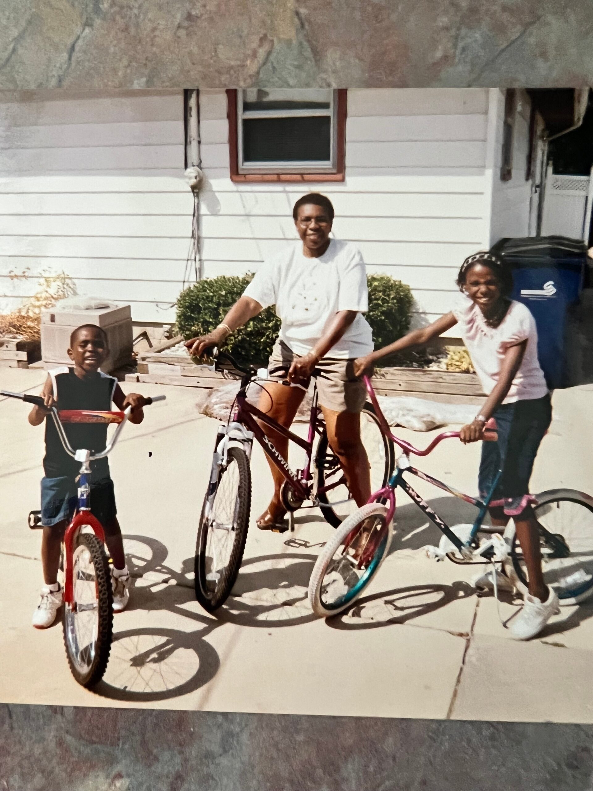 A man rides a bicycle with two kids on a sunny driveway in front of a white house with a window behind them.