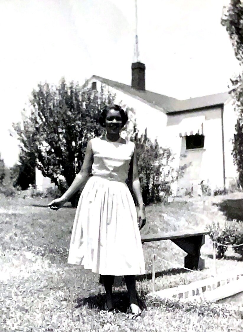 Woman in a knee-length white vintage dress posing on a lawn with a small house in the background (black-and-white photo).