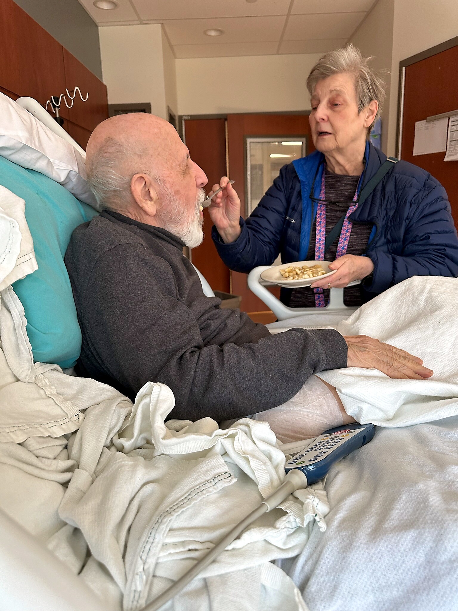 Older man with white beard sits up in a hospital bed while a caregiver feeds him with a spoon; she holds a plate of food and wears a blue jacket.