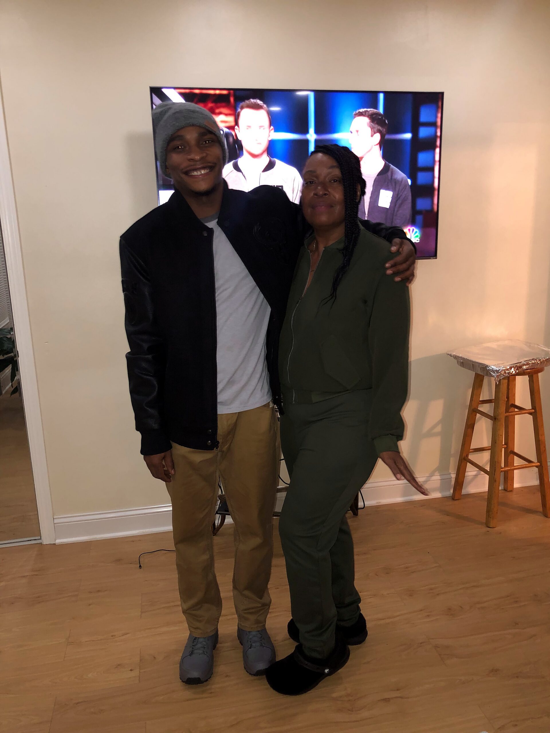 Two people pose indoors in front of a wall-mounted TV: a smiling man in a gray beanie and dark jacket beside a woman in a green outfit, with their arms around each other.