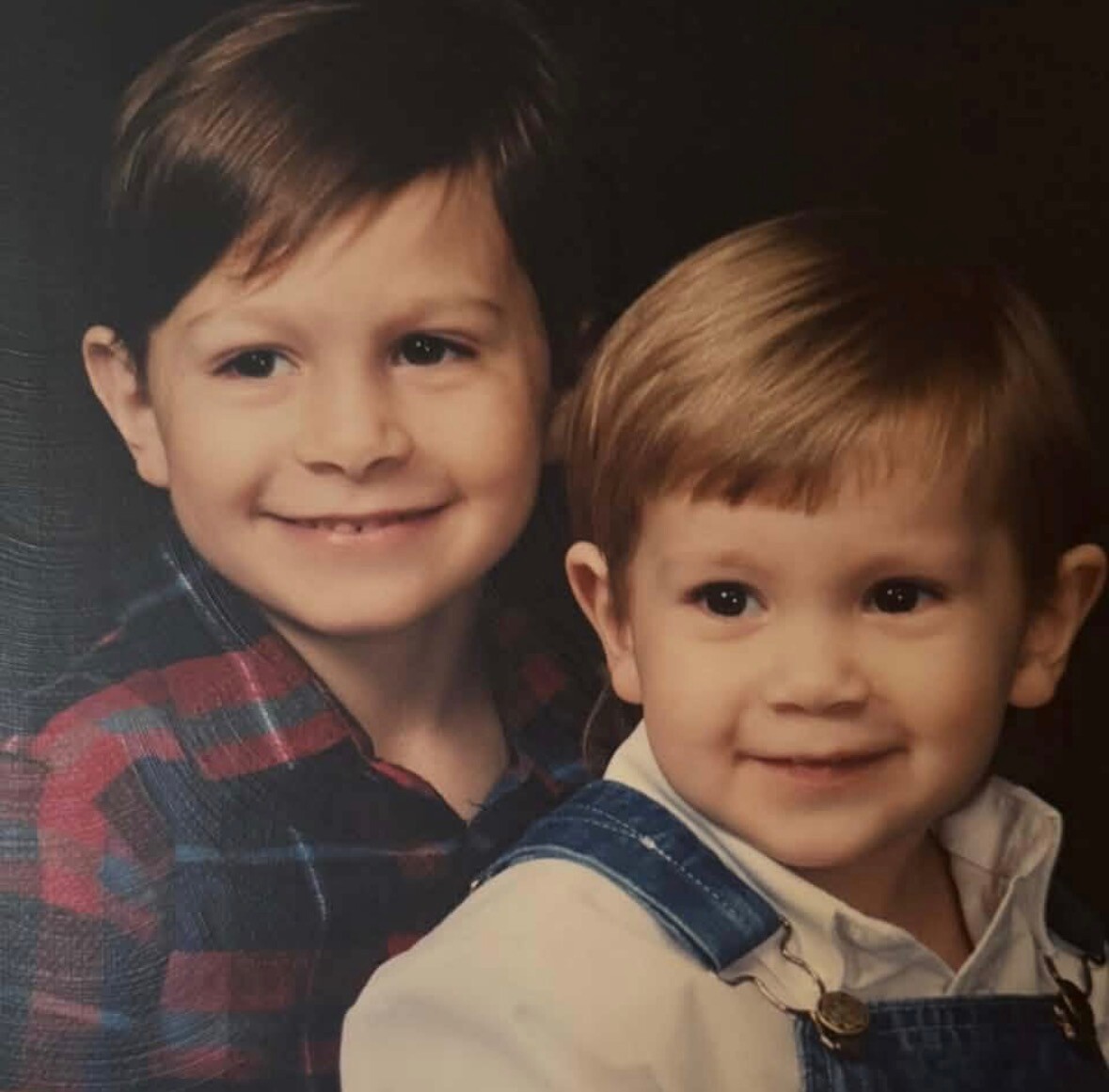 Two young boys smiling for a portrait; the older in a red plaid shirt and the younger in denim overalls with a light shirt.