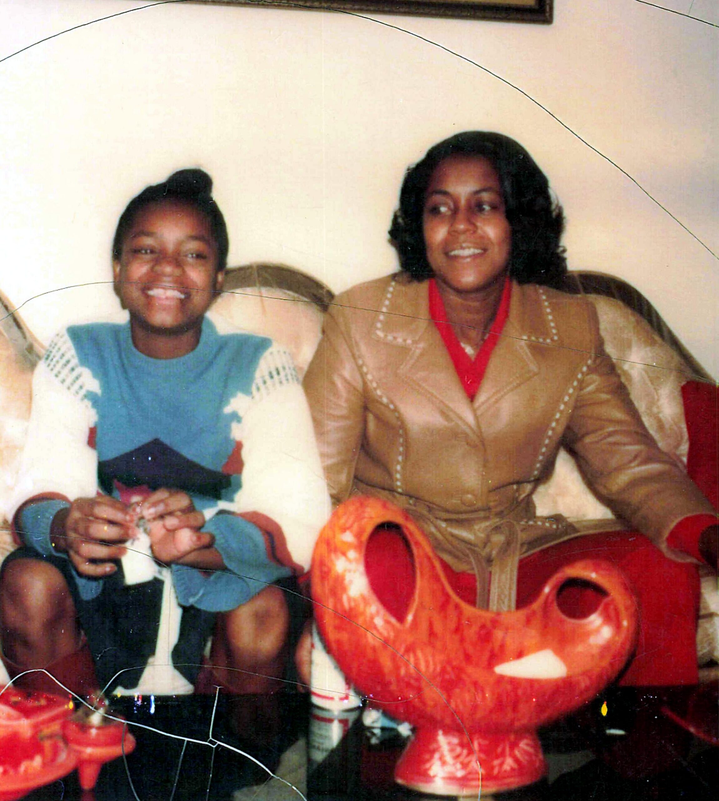 Two people sit on a couch smiling; left person in a blue and white sweater, right person in a tan jacket with a red scarf, with a red decorative dragon prop in the foreground.