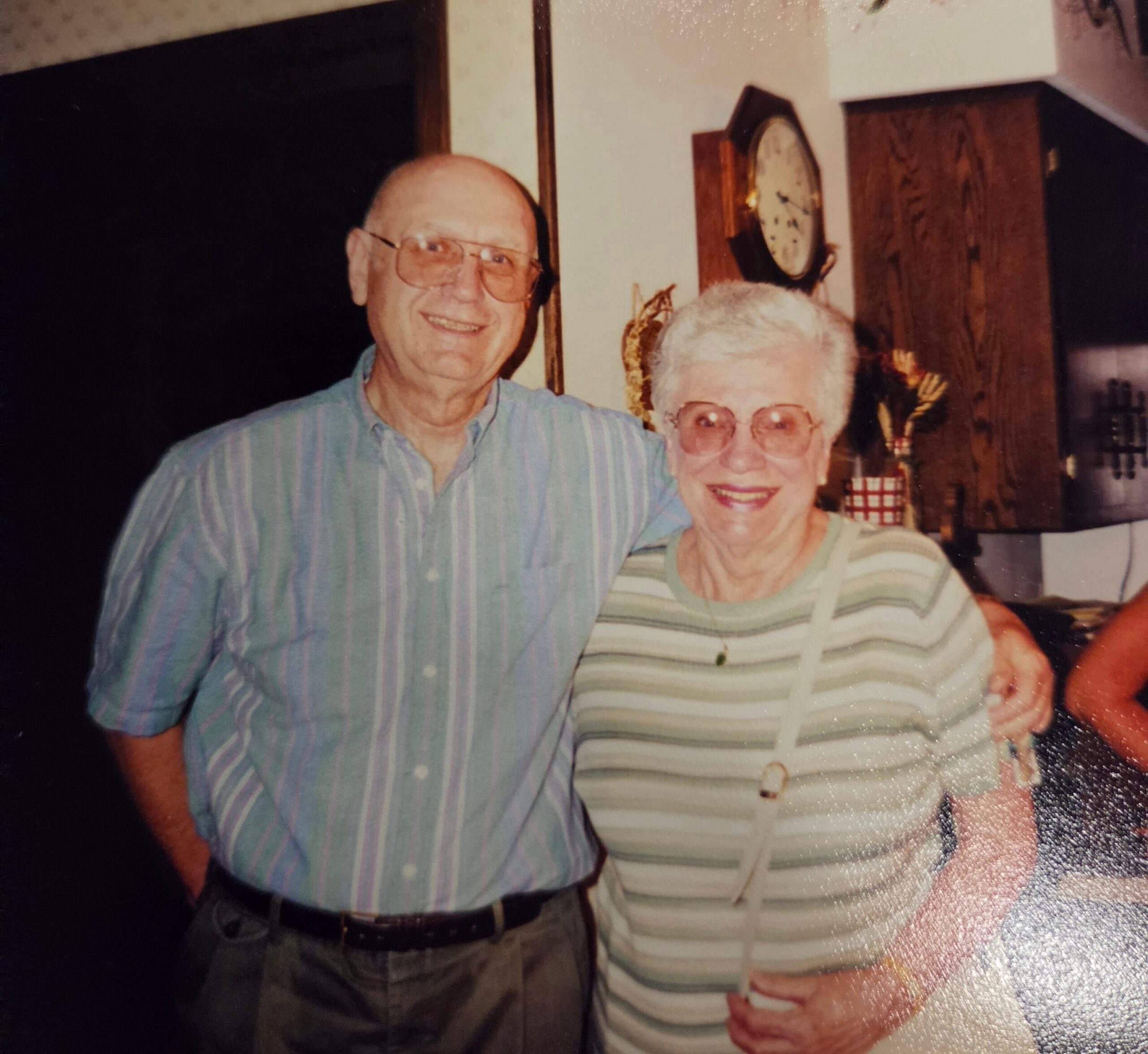 Smiling elderly couple standing close together indoors, the man in a blue striped shirt and the woman in a striped top wearing glasses.