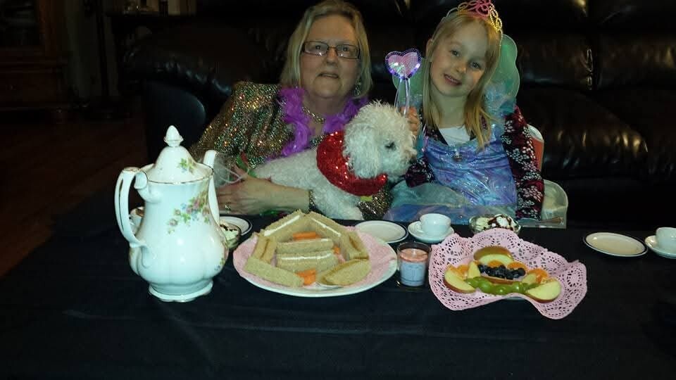 Two females in costumes sit at a tea party table with snacks, a white fluffy dog wearing a red scarf between them, all smiling.
