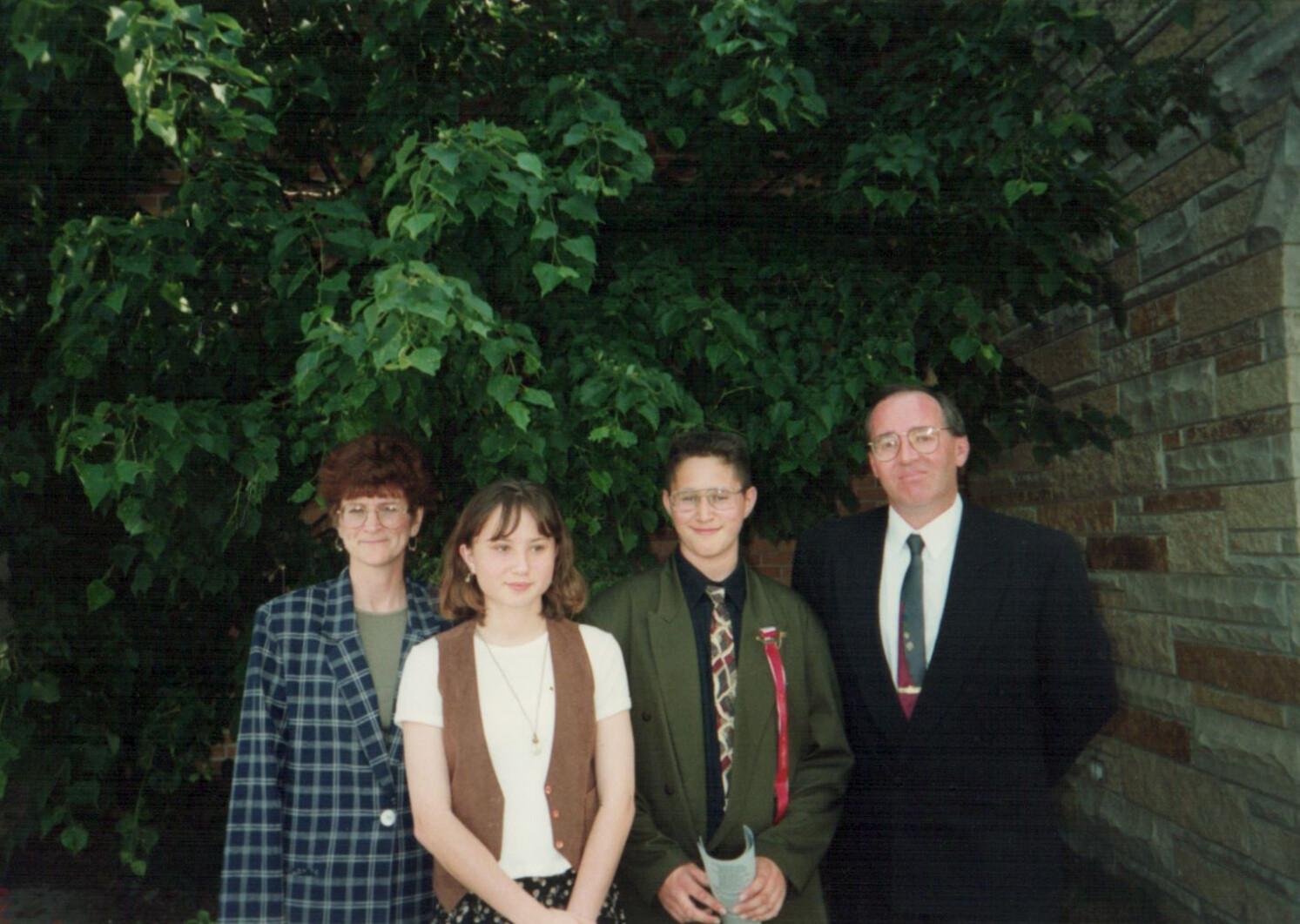 Four people stand side by side outdoors in front of lush green foliage and a stone wall.
