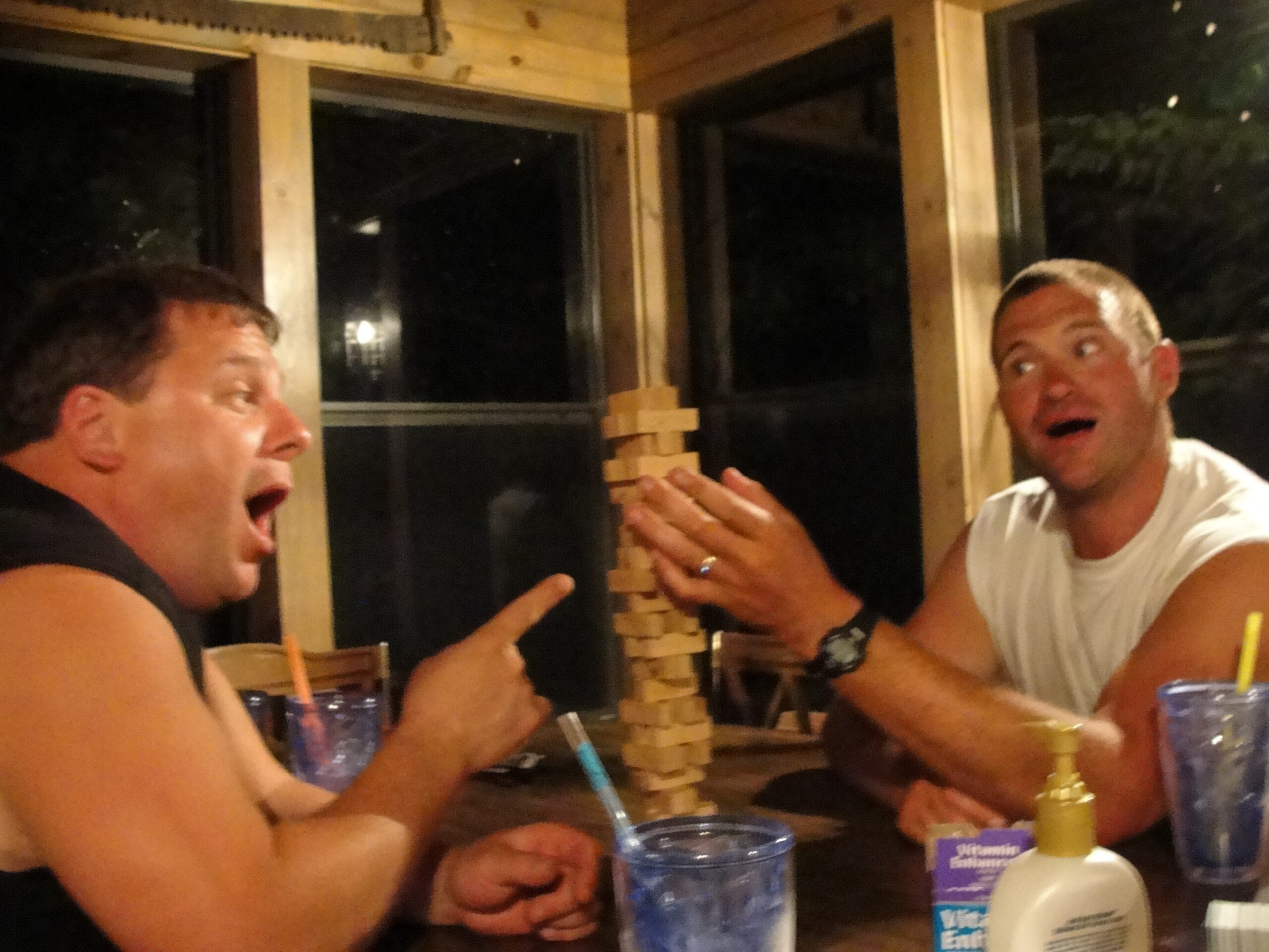 Two men at a wooden-table restaurant activity, one building a Jenga tower while the other watches with surprised expression.