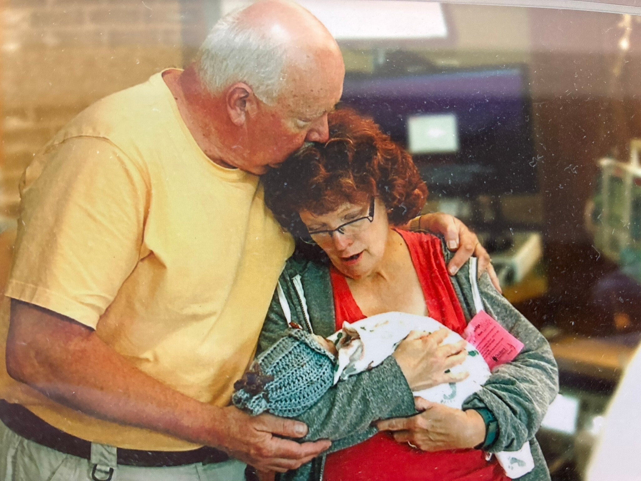 Older man with gray hair hugging a woman, their heads together in a warm embrace indoors.