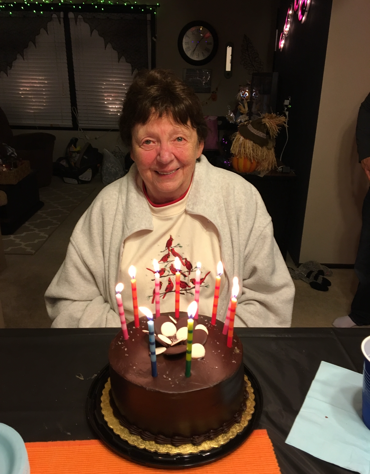 Smiling elderly woman in a light cardigan sits at a table with a chocolate birthday cake and many lit candles in front of her in a cozy room.