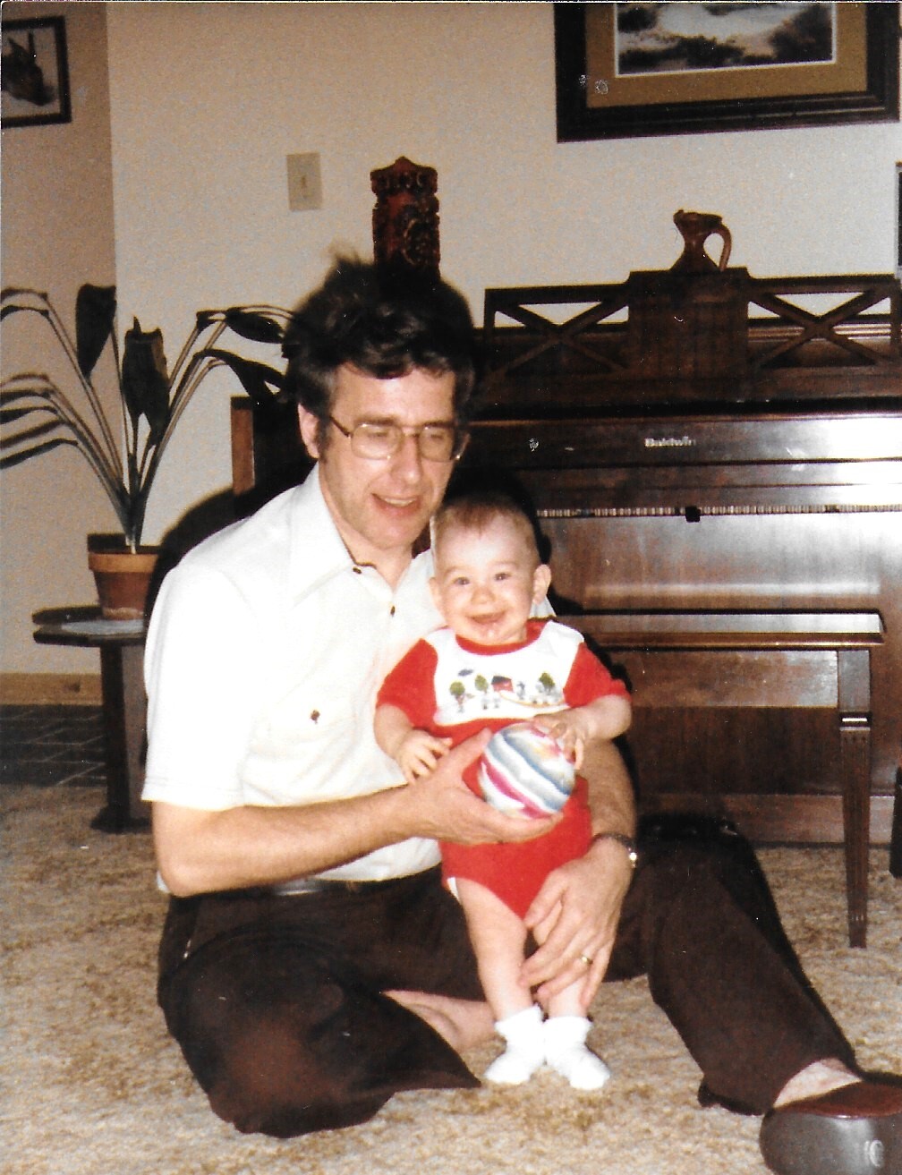 Older man with glasses sitting on the floor, holding a smiling baby in red, with a piano in the background.
