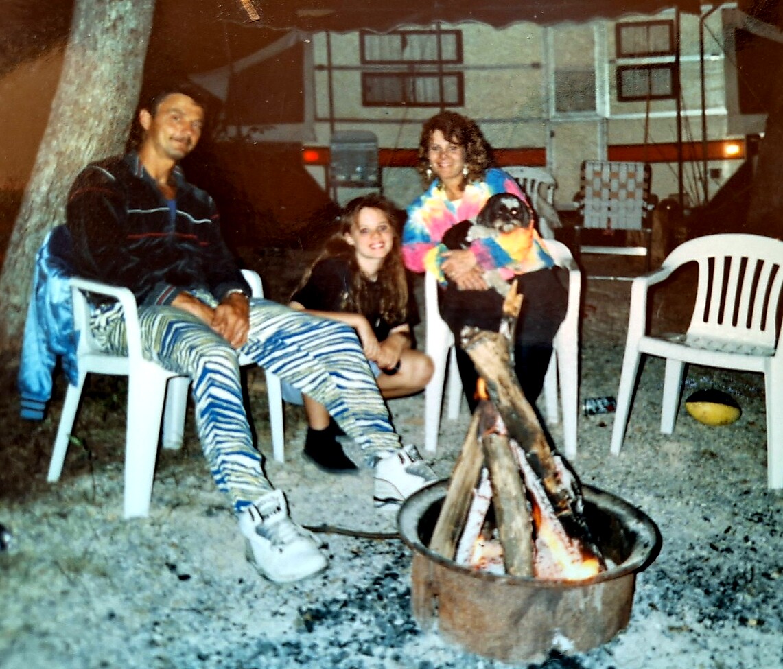 Group of four people and a small dog sitting around a campfire outdoors at night, on white plastic chairs.
