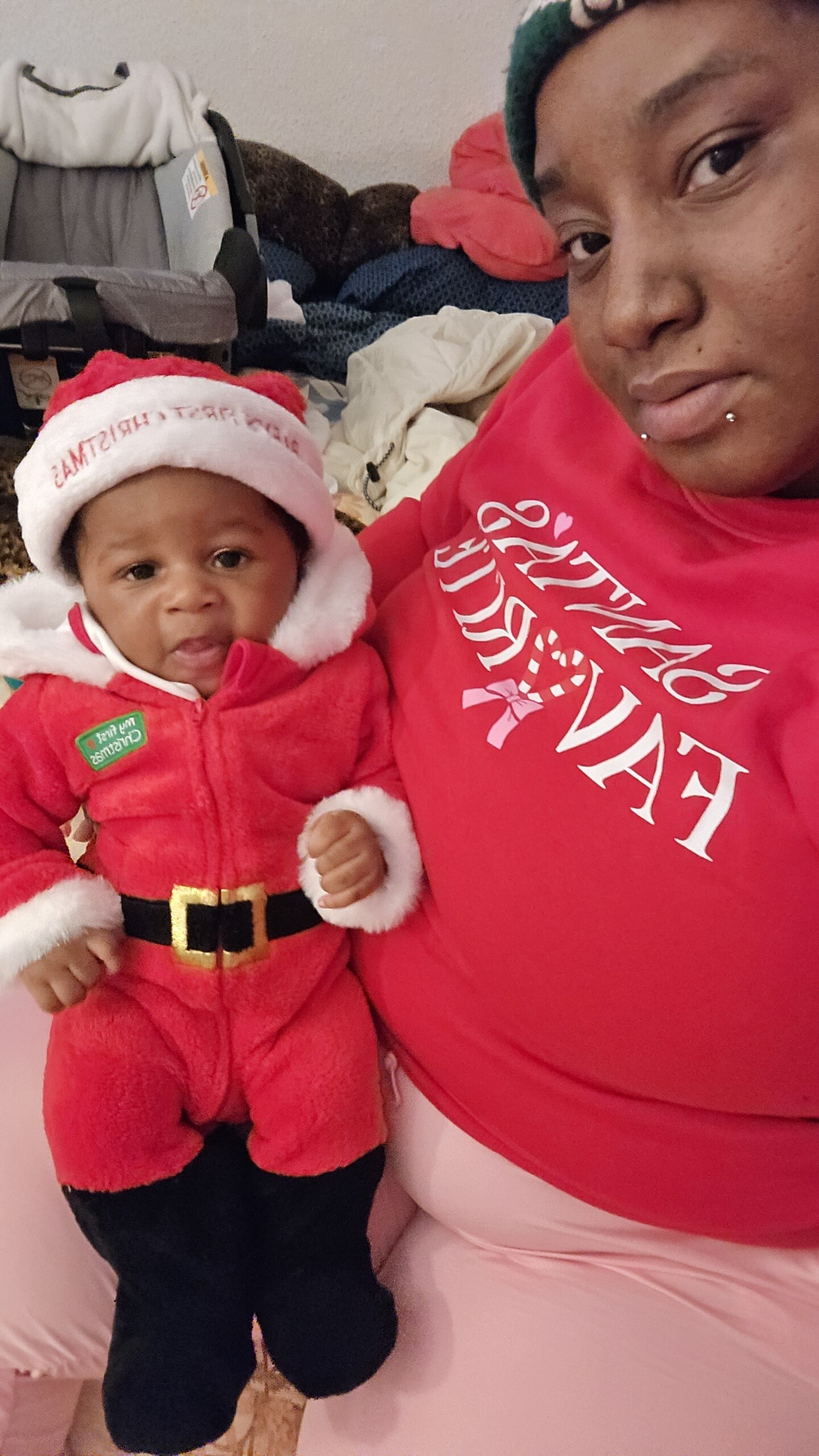 Baby in a Santa Claus outfit and hat sits beside an adult in a red shirt; festive holiday photo indoors.