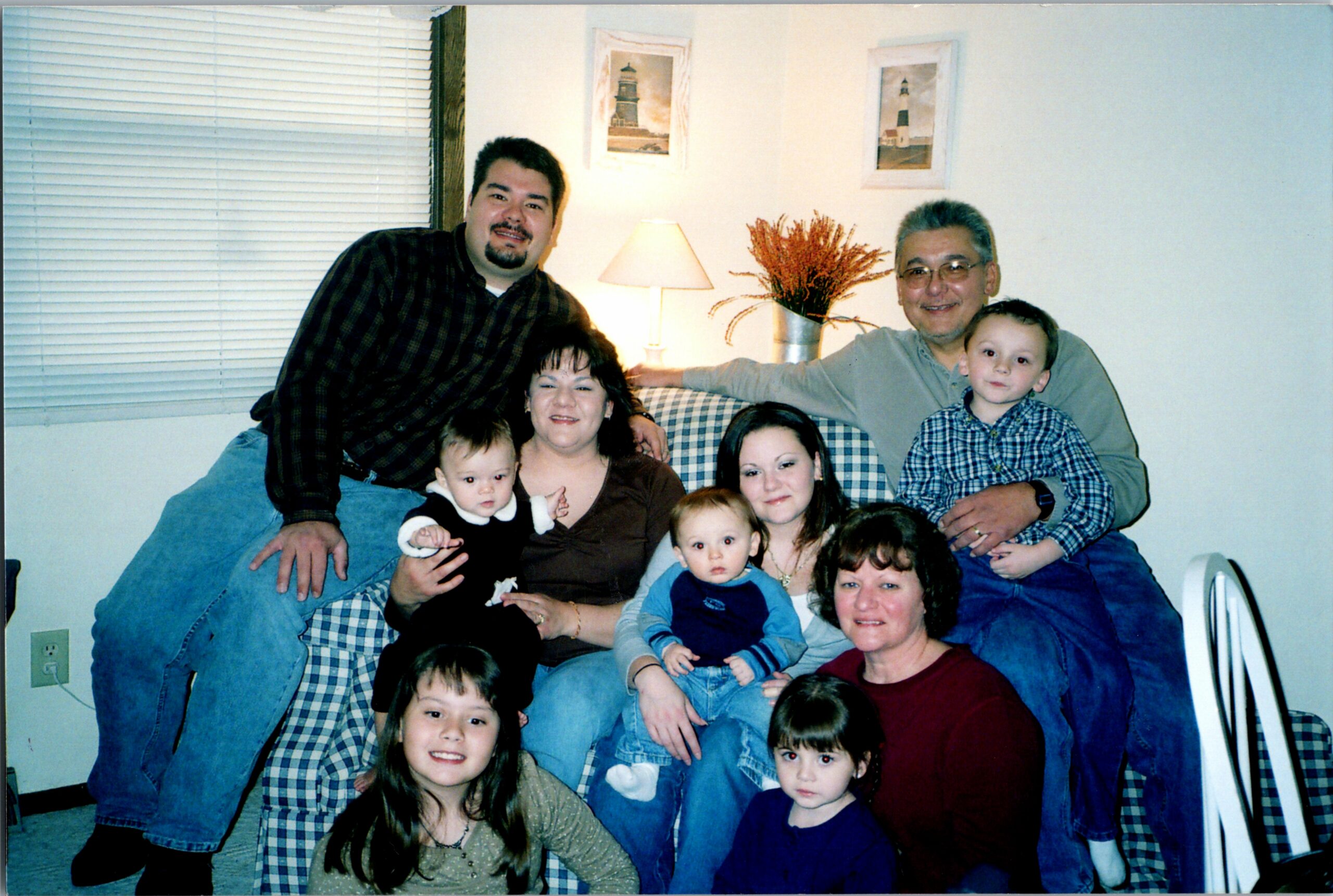 A multi-generational family sits together on a plaid couch in a living room, smiling for a group photo, with children in front and a lamp and artwork in the background.
