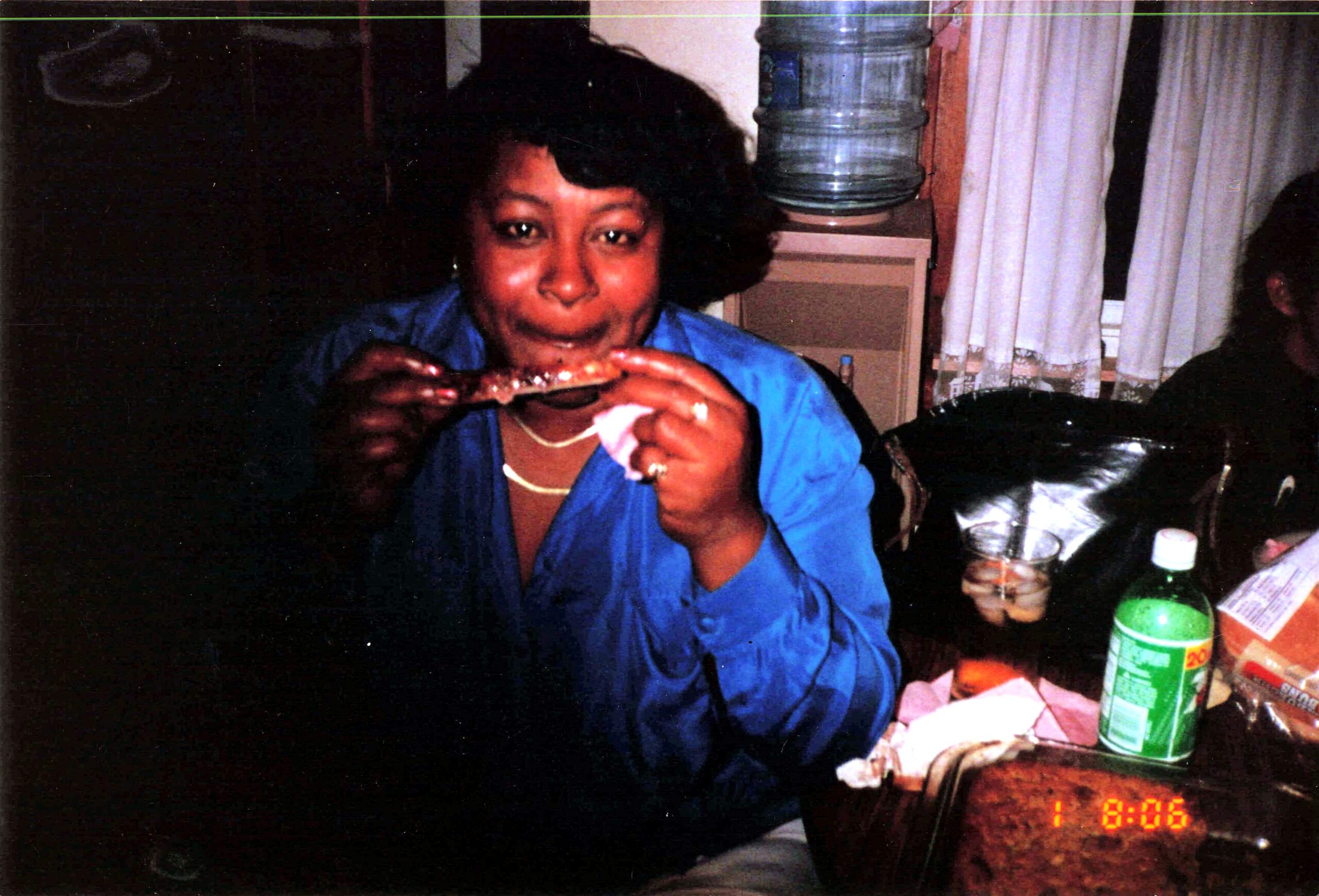 Woman in a blue blouse biting into a rib at a dim indoor gathering with snacks and bottles on the table behind her.