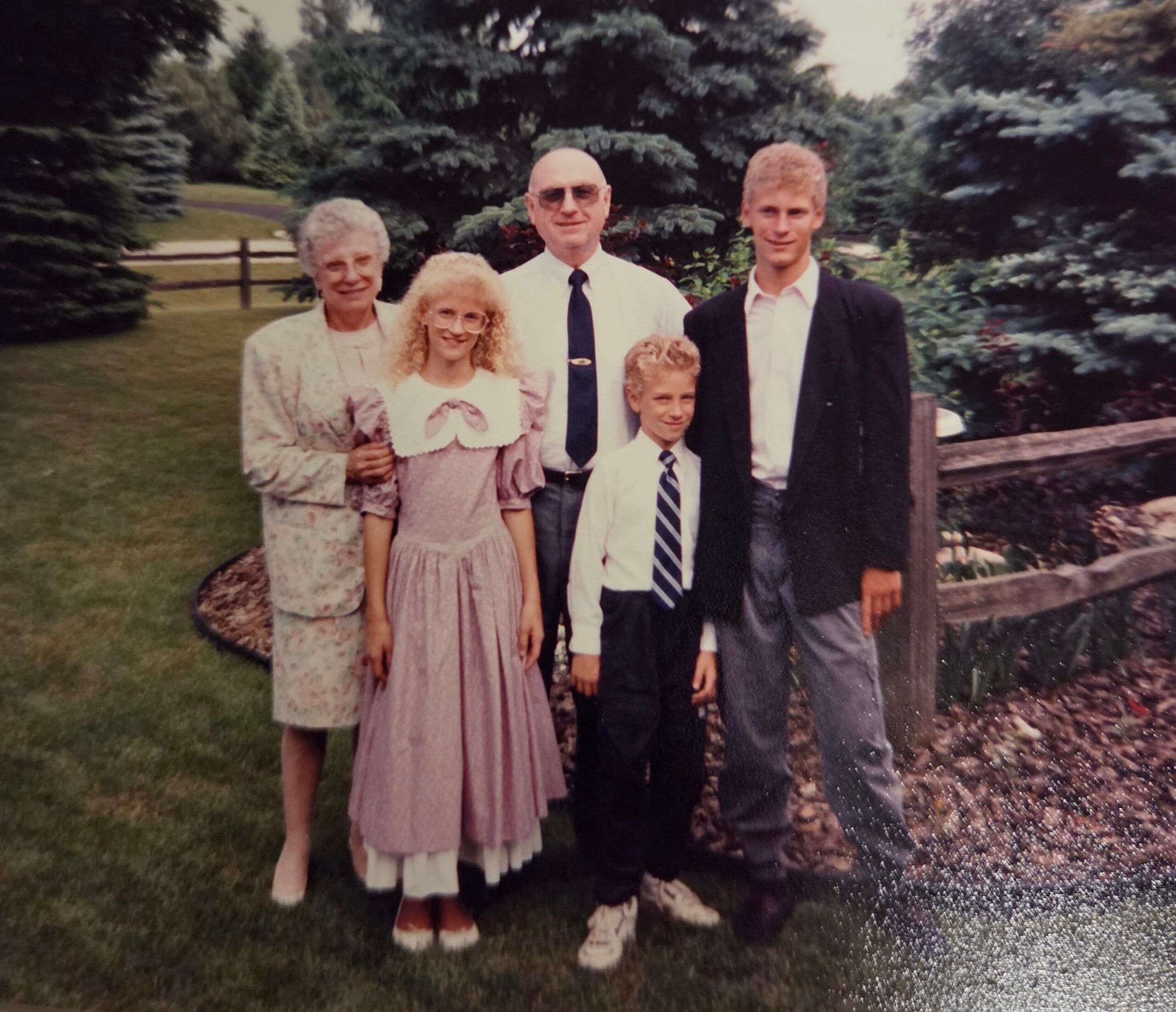 Family posing outdoors on a lawn with trees and a wooden fence in the background; two adults and two children in formal clothes.