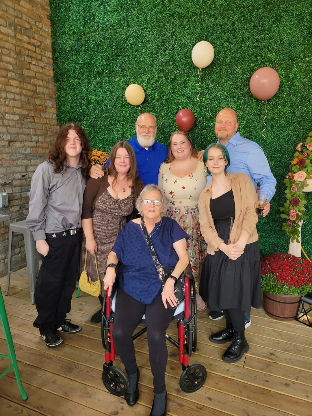 A group of eight people posing together indoors, with a woman in a red-and-black wheelchair at the front center, in front of a green hedge wall and balloons.