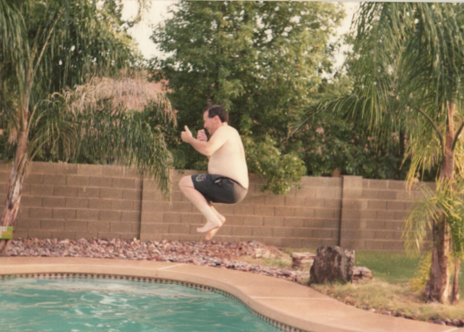 Man shirtless in black shorts leaping into a backyard pool from the deck, mid-air with trees and a brick wall behind.