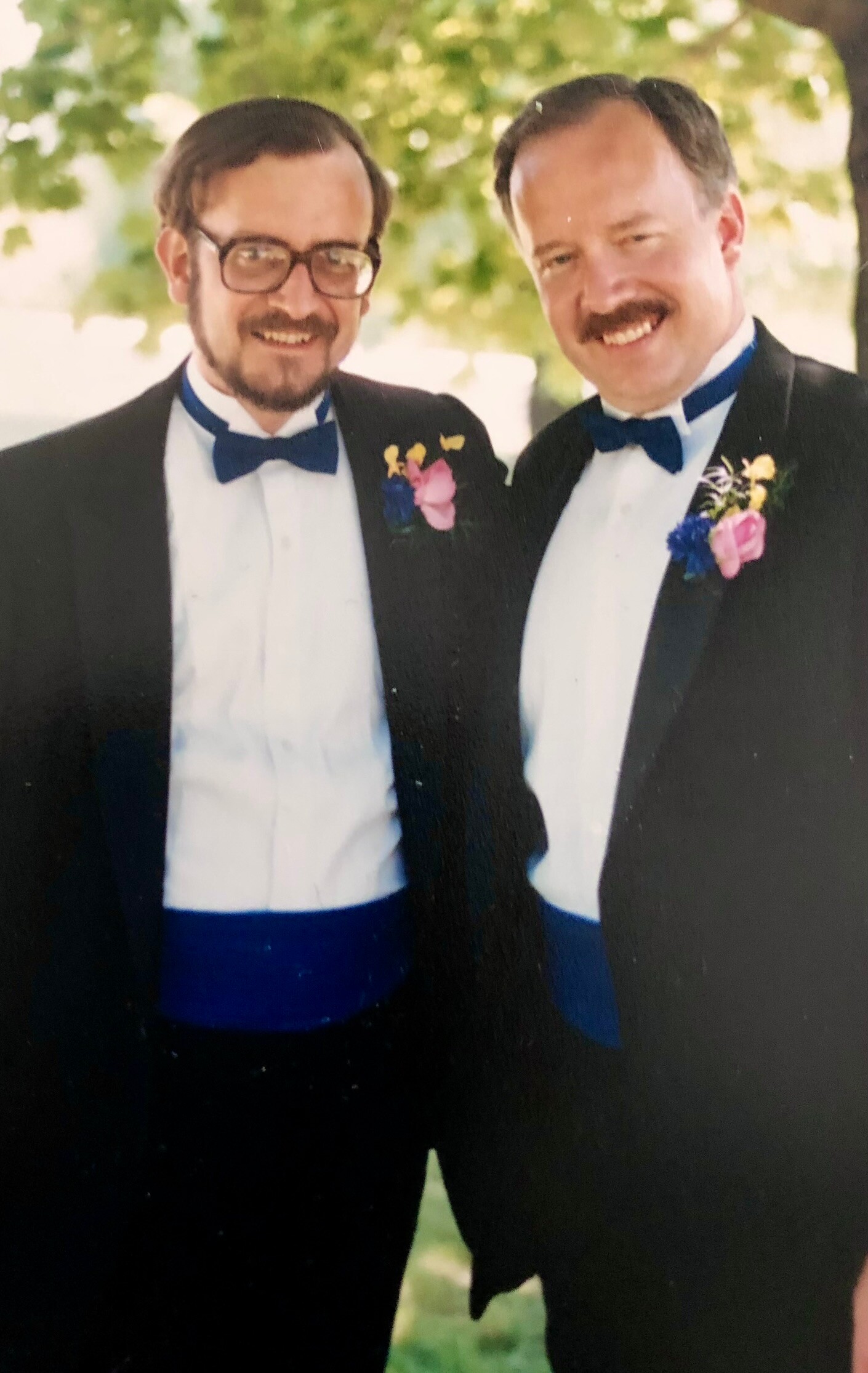 Two men in black tuxedos with blue bow ties, smiling for a photo at an outdoor wedding or formal event, each wearing a colorful boutonniere on the left lapel.