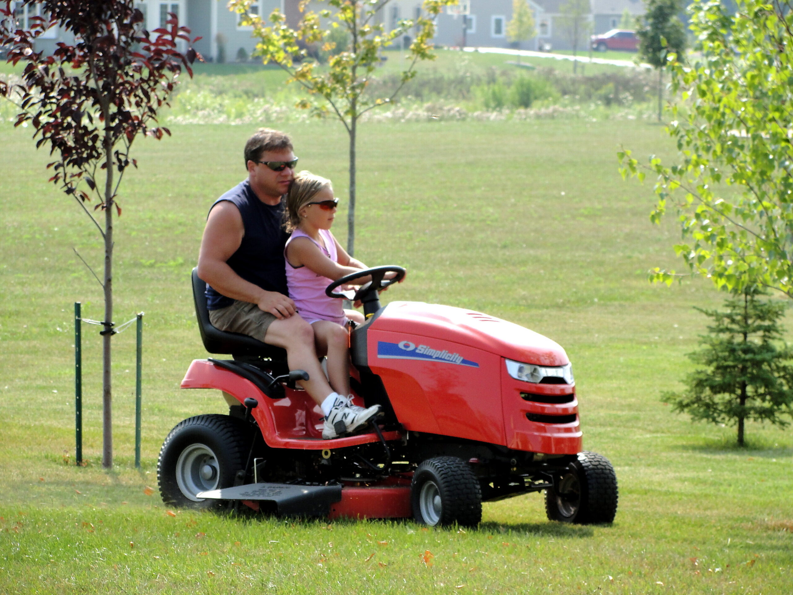 Man and young girl ride a red lawn tractor across a sunny grassy field, both wearing sunglasses and seated on the mower.