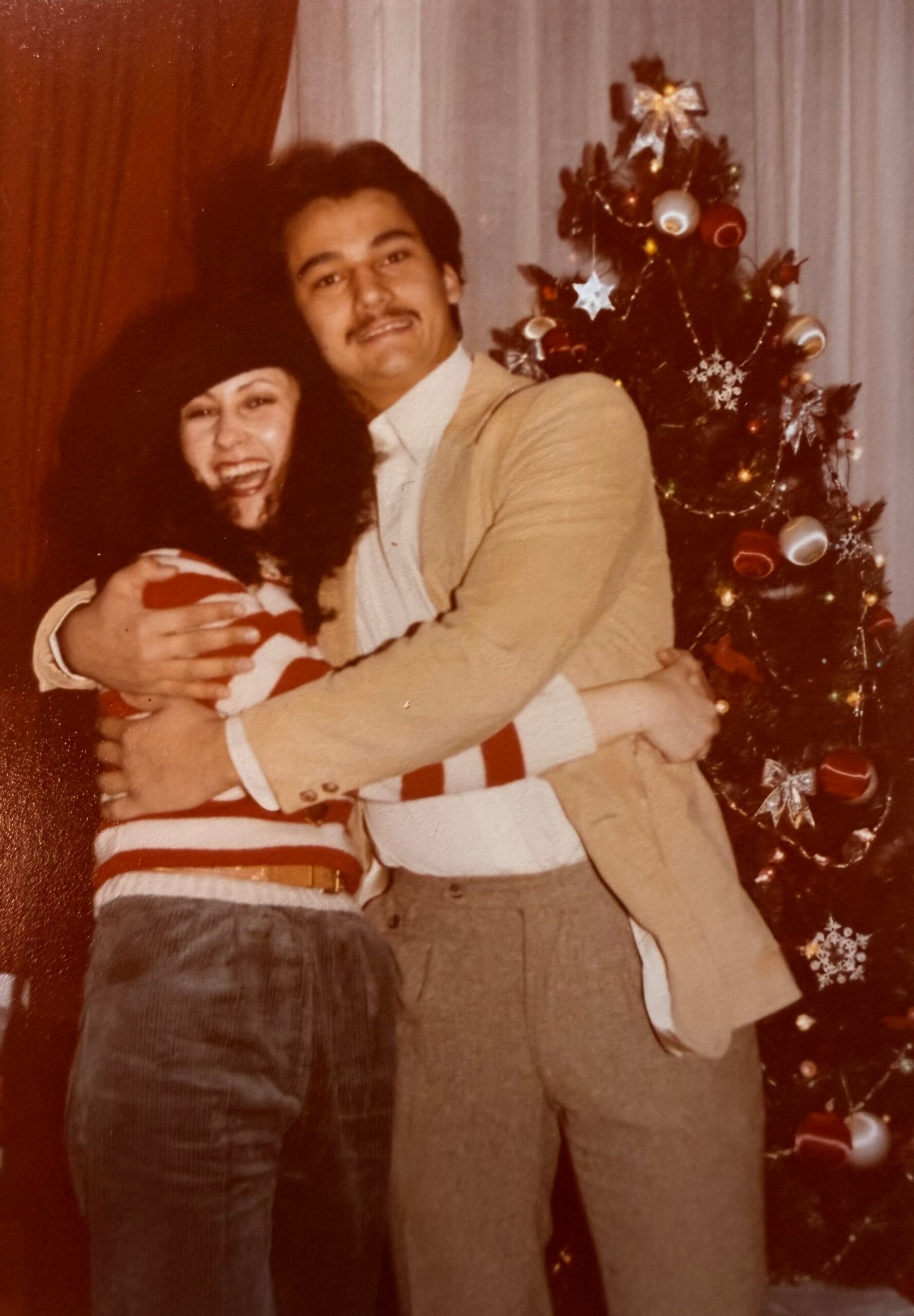 Two people hug and smile at the camera beside a decorated Christmas tree in a festive indoor setting.