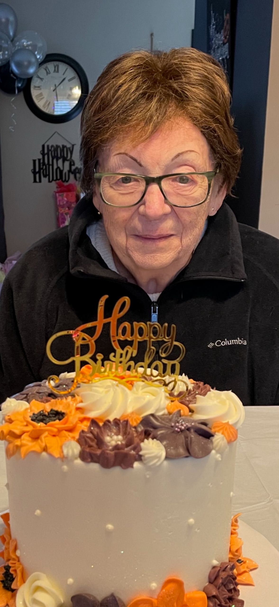 Older woman with glasses smiling at a birthday cake topped with a gold 'Happy Birthday' sign, surrounded by orange, purple, and white frosting.