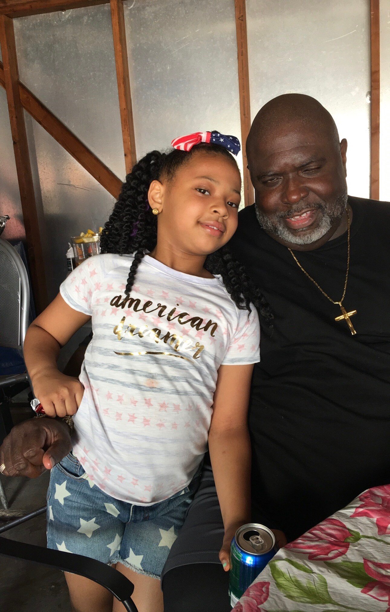 Smiling young girl with braided hair and an American flag bow posing with an older man wearing a gold cross necklace.