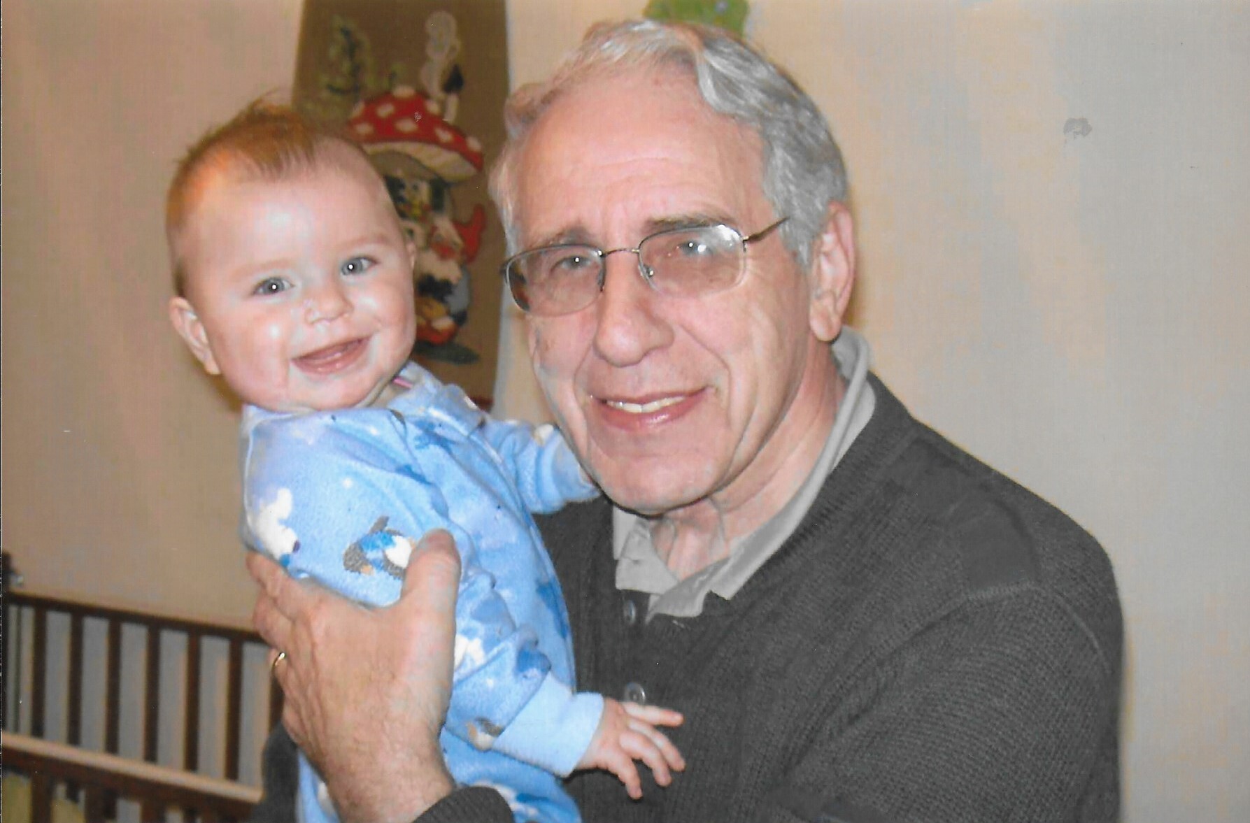 Smiling grandfather in glasses holding a baby in a blue onesie indoors.