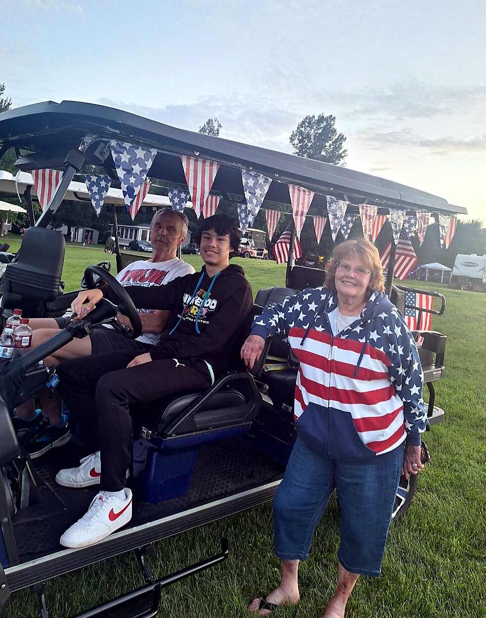 Three people in a decorated golf cart at a patriotic event; flags strung above, man driving, teen passenger, woman in a flag-themed hoodie beside them.