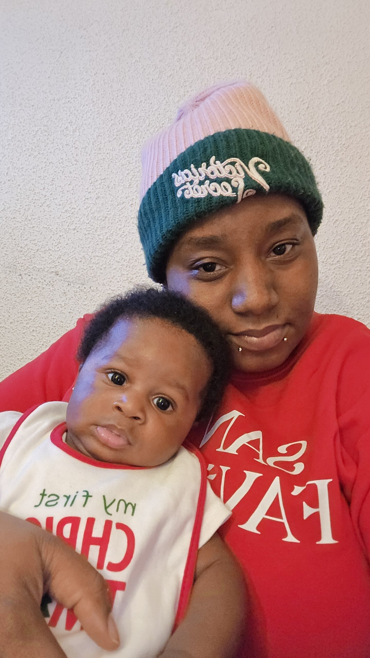 Adult wearing a pink-and-green knit beanie holds a baby in a white onesie with red trim, both looking at the camera against a light wall.